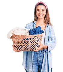 Young beautiful girl holding laundry basket looking positive and happy standing and smiling with a confident smile showing teeth - obrazy, fototapety, plakaty