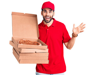 Young handsome man with beard wearing delivery uniform holding boxes with pizza celebrating victory with happy smile and winner expression with raised hands