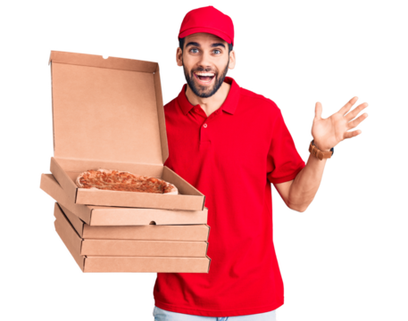 Young handsome man with beard wearing delivery uniform holding boxes with pizza celebrating victory with happy smile and winner expression with raised hands - Powered by Adobe