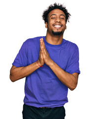 Young african american man with beard wearing casual purple t shirt praying with hands together...