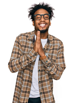 Young african american man with beard wearing casual clothes and glasses praying with hands together asking for forgiveness smiling confident.