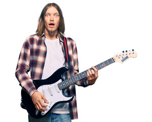 Handsome caucasian man with long hair playing electric guitar scared and amazed with open mouth for...