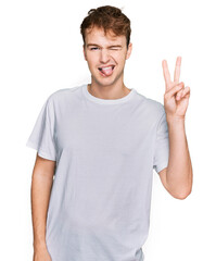 Young caucasian man wearing casual white t shirt smiling with happy face winking at the camera doing victory sign. number two.