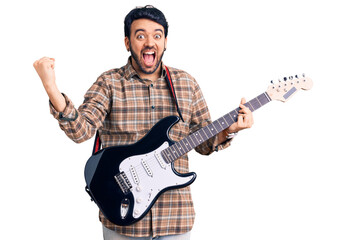Young hispanic man playing electric guitar screaming proud, celebrating victory and success very excited with raised arms