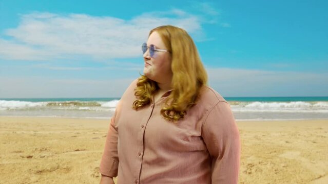 Confident curvy woman in sunglasses winking on camera on sunny beach background