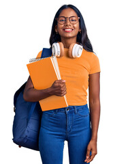 Young indian girl holding student backpack and books looking positive and happy standing and smiling with a confident smile showing teeth - obrazy, fototapety, plakaty