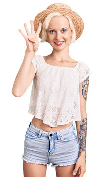 Young blonde woman with tattoo wearing summer hat showing and pointing up with fingers number four while smiling confident and happy.