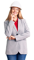 Beautiful young woman wearing architect hardhat looking confident at the camera with smile with crossed arms and hand raised on chin. thinking positive.