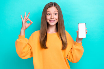 Photo of cute pretty nice child girl wear orange sweatshirt showing okey sign hold phone screen ad...