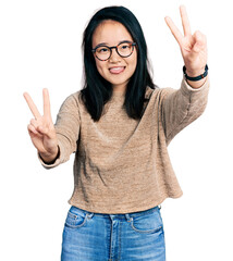 Young chinese woman wearing casual sweater and glasses smiling with tongue out showing fingers of both hands doing victory sign. number two.