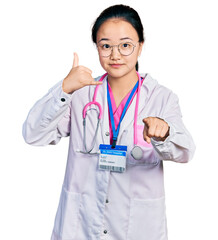 Young chinese woman wearing doctor uniform and stethoscope smiling doing talking on the telephone gesture and pointing to you. call me.