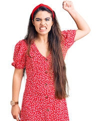 Brunette teenager girl wearing summer dress angry and mad raising fist frustrated and furious while shouting with anger. rage and aggressive concept.