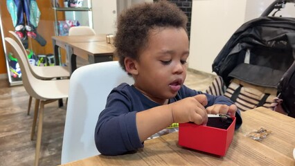 Two year-old black baby unboxing and playing with his new toy car inside a cafeteria seated next to mum - Powered by Adobe