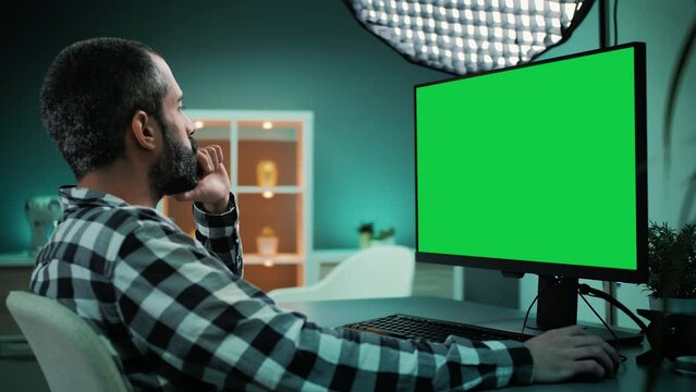 Slow motion shot over the shoulder of a handsome and focused young freelancer, copywriter, designer, editor or content creator working on desktop computer with chroma key green screen mock up display.
