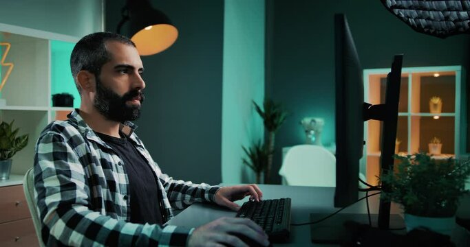 Focused young man typing on desktop keyboard doing research. Handsome male freelancer with beard working at his desk, on a modern home office studio with nice teal and orange lights in the background.
