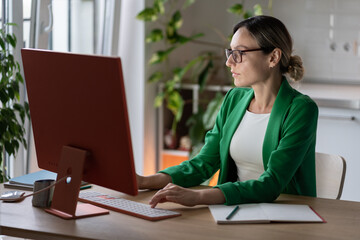 Focused freelancer making future project types information on computer. Young businesswoman wearing glasses learns important details about project and works in cozy home office with green houseplants