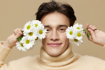 Obraz na płótnie Canvas A handsome man with light white skin, with dark, short, short hair combed back, in a beige turtleneck with a high collar stands on a dark beige background with white daisies near his face.Close-up