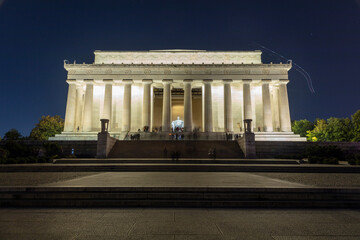 Tourists visit the Lincoln Memorial in Washington, D.C. on an autumn night. Long exposure wide shot from the east side of the building. The lights of a passing airplan are seen in the sky..