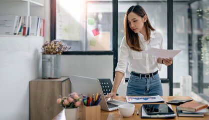 The confident asian businesswoman working on laptop compute, holding paperwork and standing at office desk.