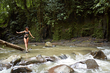 A young, slender girl poses on a tree trunk that has fallen into the river in the middle of a dense jungle