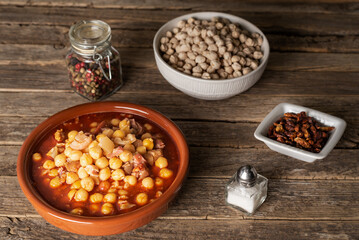 Clay dish with tripe and chickpeas, along with the ingredients used in its preparation, on an old wooden table.