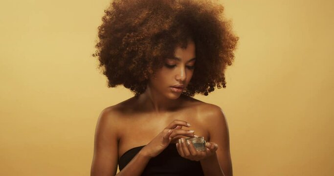 Young beautiful african american girl with perfect skin holding cosmetic jar and applying face cream. Concept of female beauty