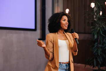 African businesswoman talking to the public over the microphone, giving the lecture. - 549176279