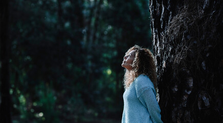 Thoughtful woman standing against a trunk tree in the forest looking up. Concept of environment and...