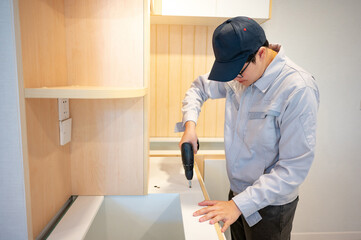 Home renovation or House remodeling concept. Asian male furniture assembler or Interior construction worker man using electric drill screwdriver installing wooden counter and cabinet of new kitchen.