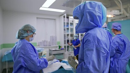 Smiling male doctor wearing highly-protective suit stands in surgery room. Female nurse helps the surgeon to put on latex gloves.