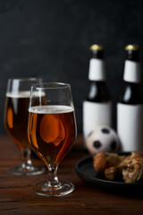Two glasses with beer, bottles, snacks and soccer ball on wooden table.