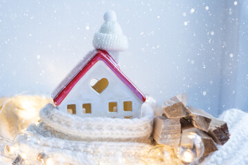 Cozy house is wrapped in a hat and scarf in a snowstorm -window sill decor. Winter, snow - home insulation, protection from cold and bad weather, room heating system. Festive mood, Christmas, New Year