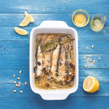 Recipe of marinated  sardines or Sarde in saor, a typical dish from the Venice, Italy  with white onions marinated with wine vinegar, raisins and pine nuts. Blue marine wooden background. Square crop