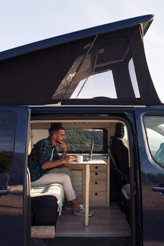 young man sitting in his camper van working on his laptop, concept of freedom and digital nomad lifestyle
