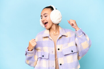 Young Arab woman wearing winter muffs isolated on blue background celebrating a victory