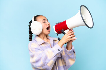 Young Arab woman wearing winter muffs isolated on blue background shouting through a megaphone