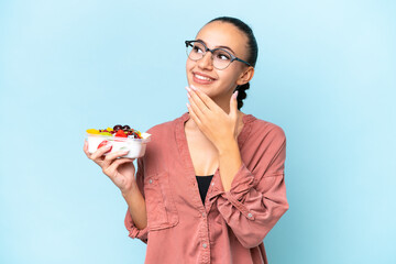 Young Arab woman holding a bowl of fruit isolated on blue background looking up while smiling