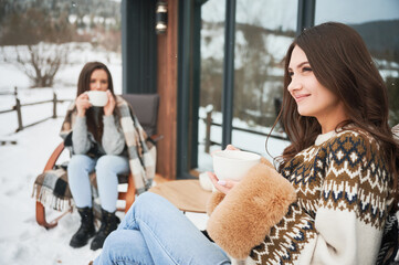Young women enjoying winter weekends on terrace of contemporary barn house in the mountains. Two girls sitting on chairs and drinking hot tea.
