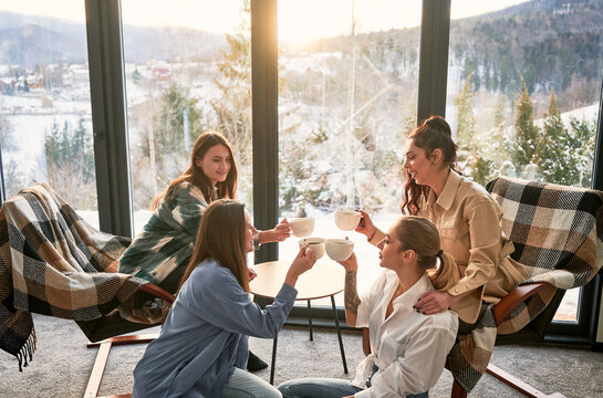 Young women enjoying winter weekends inside contemporary barn house. Four girls having fun and clinking cups of hot tea near panoramic windows at sunset.