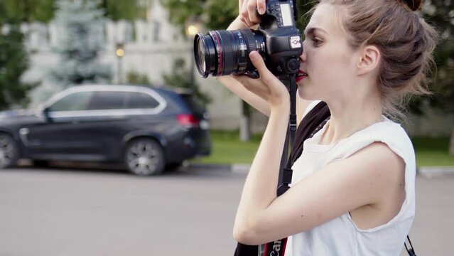 professional photographer in white T-shirt and with bun on her head holds black camera in hands and takes picture. woman take photos and adjusts camera, look into viewfinder
