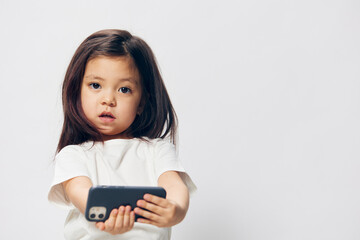 a cute little preschool girl stands on a white background in a white T-shirt and takes pictures of herself on her phone, smiling happily at the camera. The theme of children's happiness