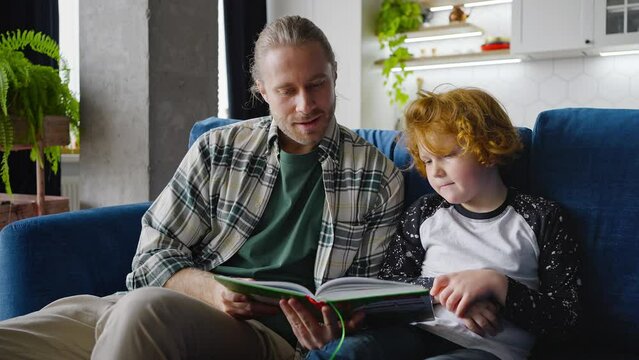 Bearded father reads book to little son sitting on blue sofa