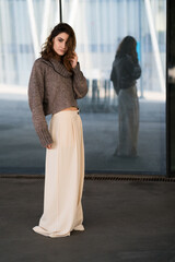 Portrait of a young adult woman standing in front of a reflective glass looking at camera wearing fashionable pants