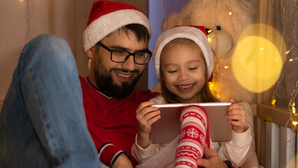 Happy family, father and little girl in red santa hats use digital tablet and laugh sitting in house bed. New Years Christmas holiday time.