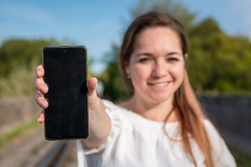 Mujer joven sonriente mostrando su celular con la pantalla apagada. Fotografía con enfoque...