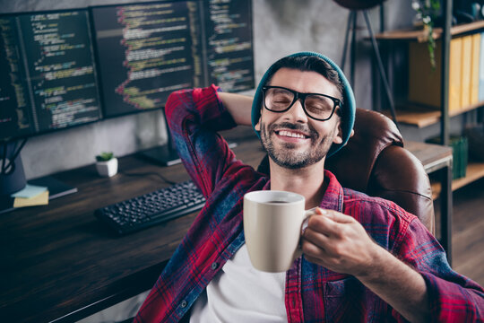 Photo of thoughtful funny coder dressed hat glasses smiling arm behind head closed eyes drinking tea indoors workplace workstation loft