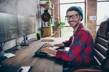Photo of cheerful positive coder dressed hat glasses smiling writing system code indoors workplace workstation loft