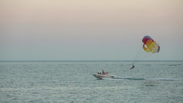Boat pulling a parachute over the blue sea at beautiful sunset. Motorboat towing a parachute with tourists along the sea against the horizon. Parasailing in calm still water. Extreme sports aerial