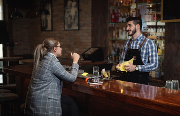 Young business woman in a formal wear sitting in a cafeteria at the bar after work and flirts with a bartender polishing glasses. Freelancer female talking to a waiter in the restaurant after work. 