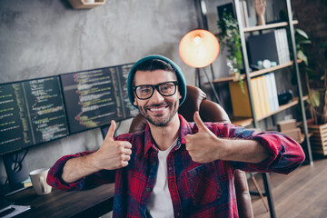 Photo of happy positive freelancer wear hat glasses showing two thumbs up smiling indoors workplace...